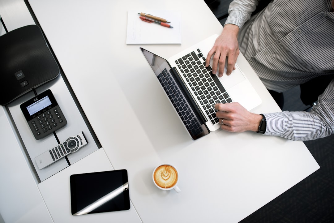 Working professional sits at a white desk with laptop, coffee, tablet, and notebook, wearing a striped shirt and watch as he works with pens and calculator nearby.