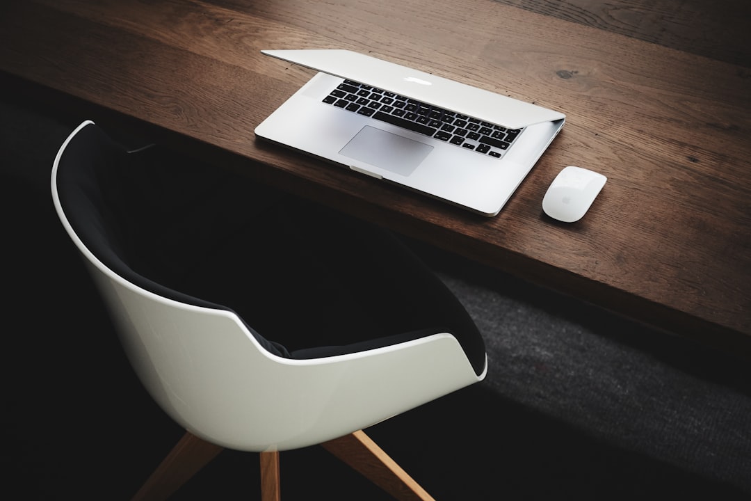 Contemporary office setup with sleek white laptop, minimalist chair, and wooden desk in dark tones, perfect for productivity and comfort in a modern workspace setting.