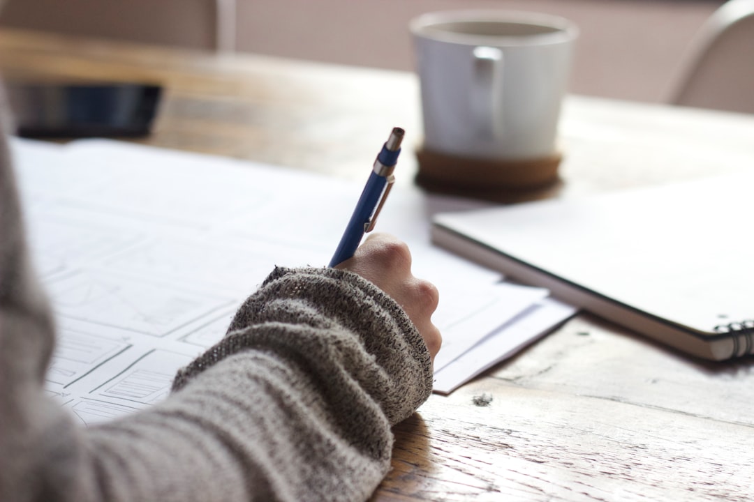A student prepares for a study session with a pen over papers on a wooden desk surrounded by a notebook and a cup of coffee in a cozy home office setting.