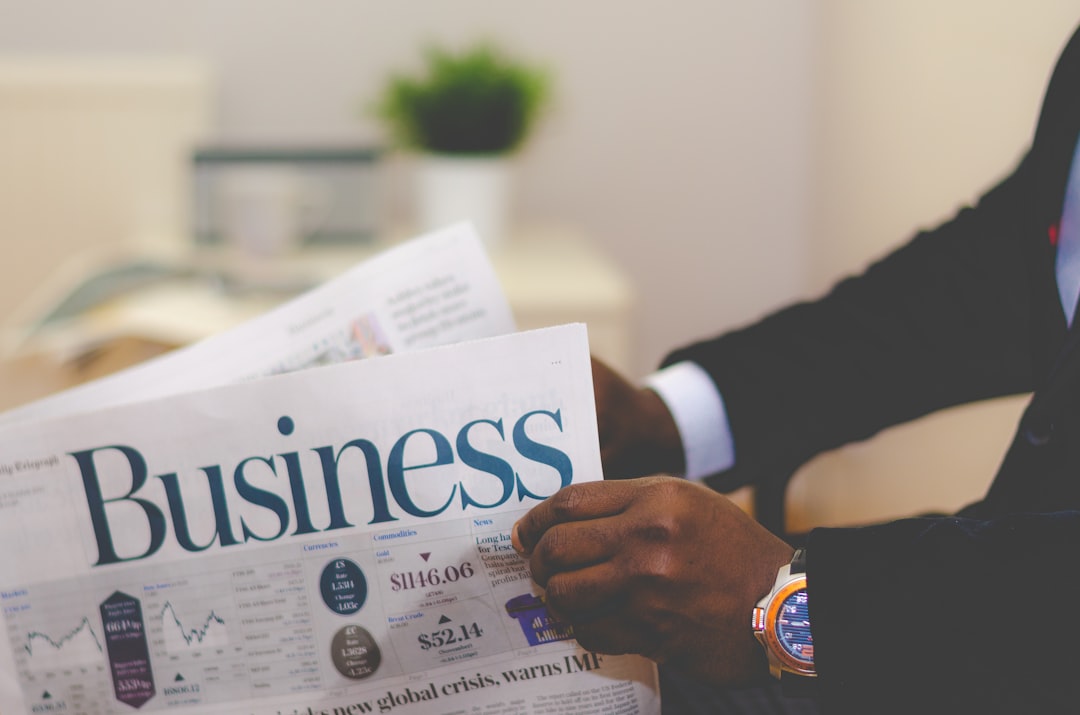 Executive wearing a watch, reading a business newspaper with financial charts and headlines in a professional office setting amidst a blurred background.