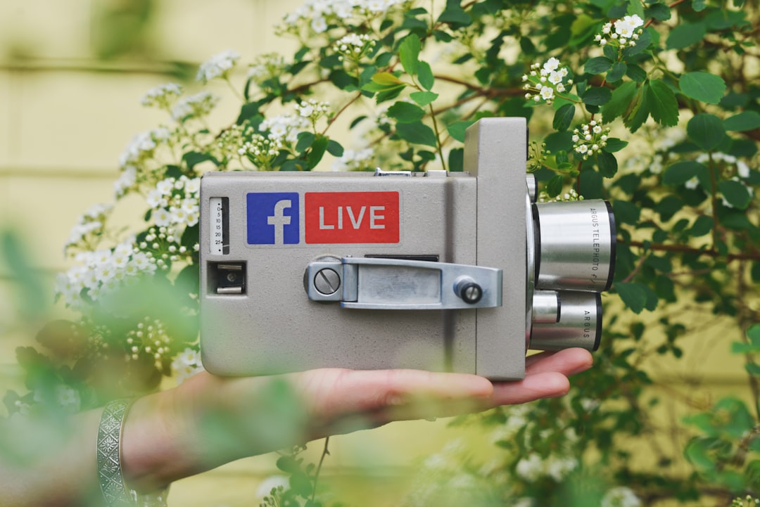 Person capturing outdoor Facebook Live broadcast using a vintage camera surrounded by lush greenery and white flowers, evoking a retro feel.