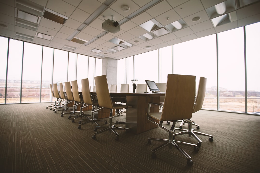 Modern conference room with large windows, wooden table, and modern office chairs, offering a bright professional meeting space with a city view background and laptops ready.