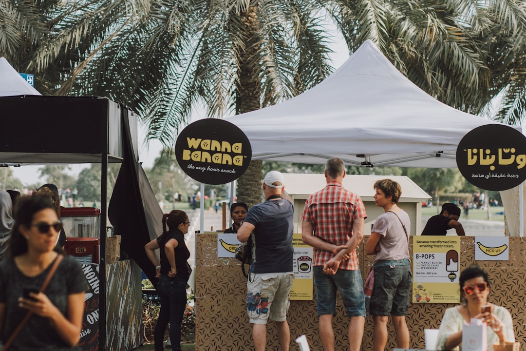 Outdoor food stall 'Wonka Bananas' under white tents, surrounded by palm trees, lively customers, and casual attendees on a sunny day, enjoying banana-based treats.